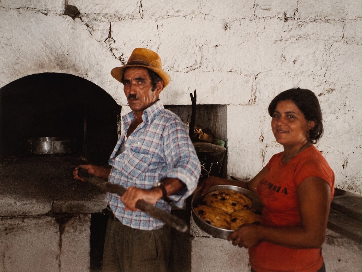 A man drawing a tray from a village fourno with a wooden peel while a woman holds a tray of freshly baked bread